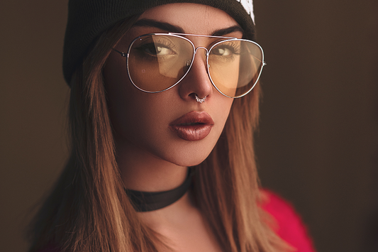 A moody image of a woman wearing orange glasses with her septum pierced