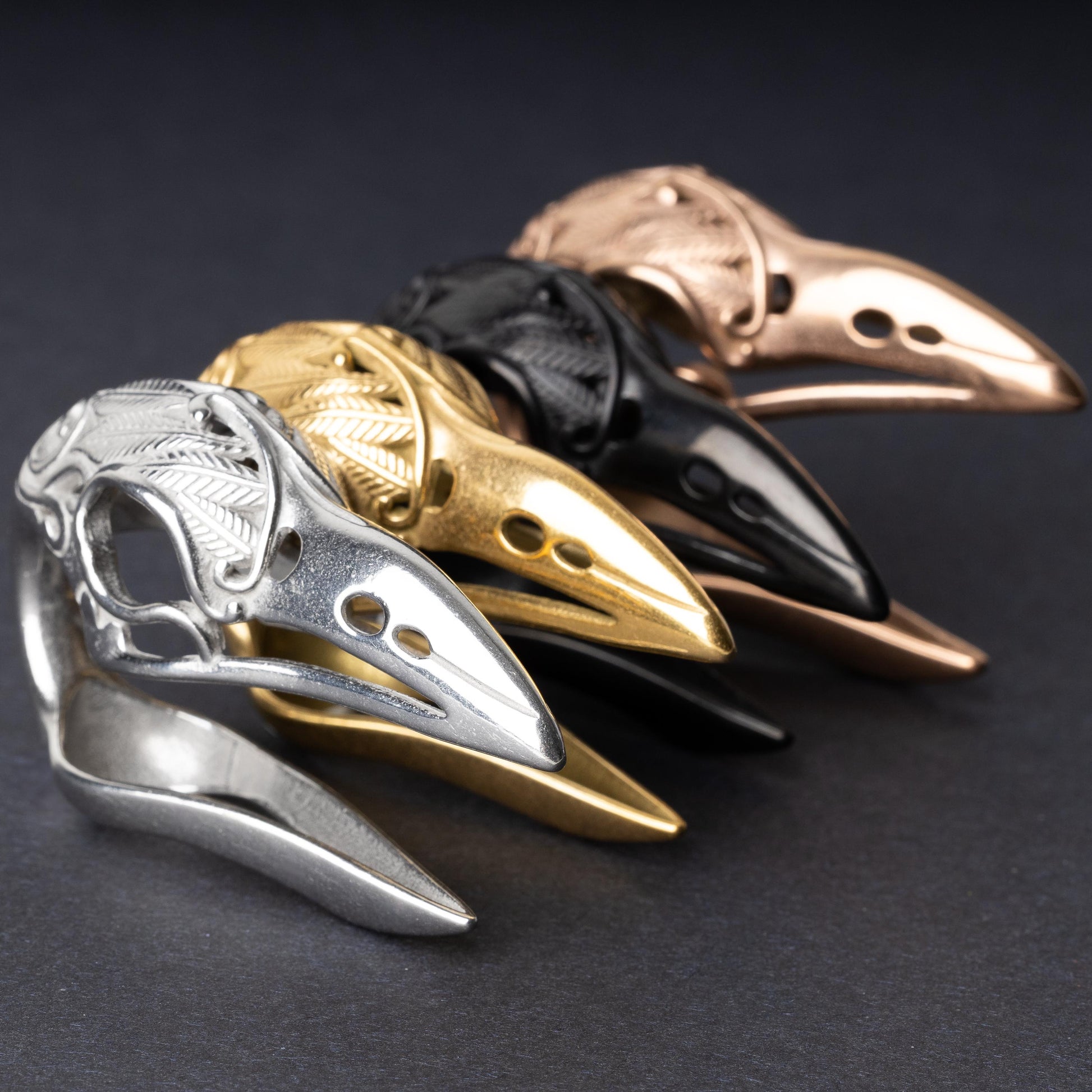 Raven skull ear hangers in silver, gold, black, and rose gold finishes, showcasing intricate feather engravings and polished details, displayed on a dark background.