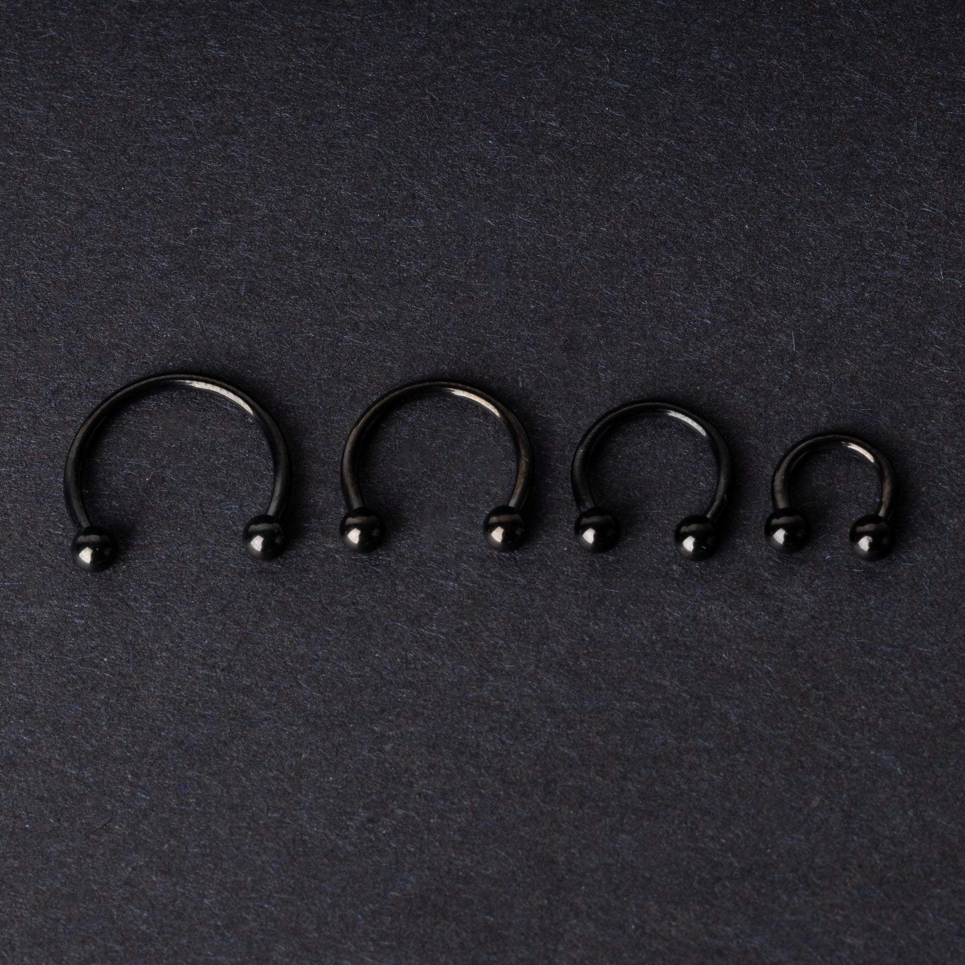 four G23 titanium black horseshoe rings with ball ends in varying sizes, displayed on a dark background showcasing their polished finish.