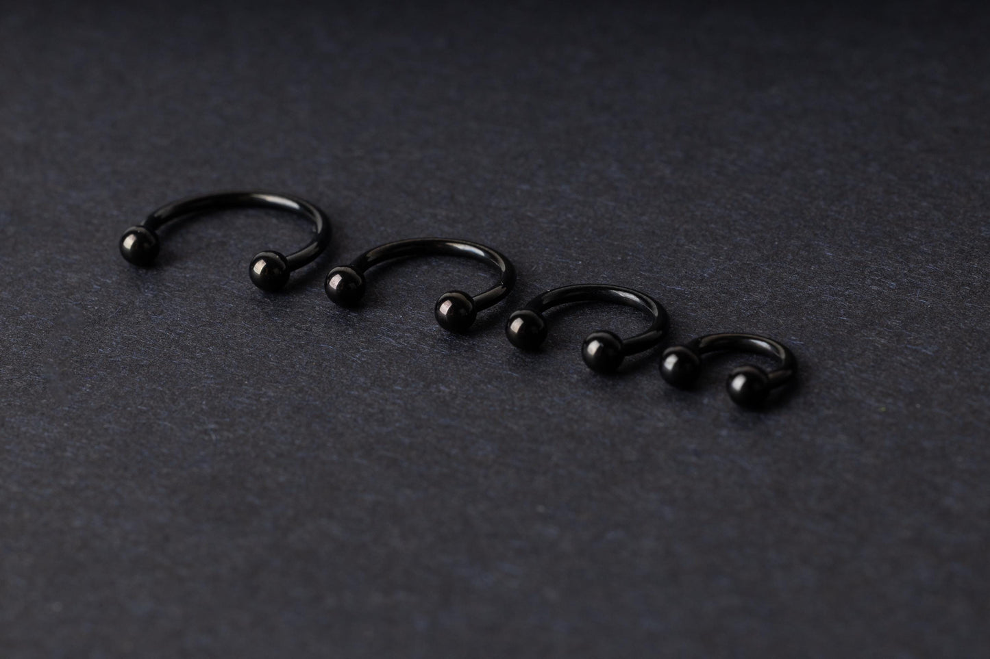 four G23 titanium black horseshoe rings with ball ends in varying sizes, displayed on a dark background showcasing their polished finish.