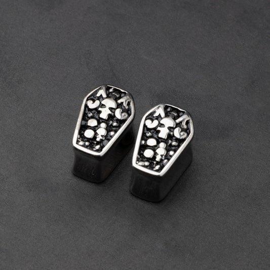 A pair of coffin-shaped stainless steel ear plugs with intricate skeleton designs, displayed on a dark background.