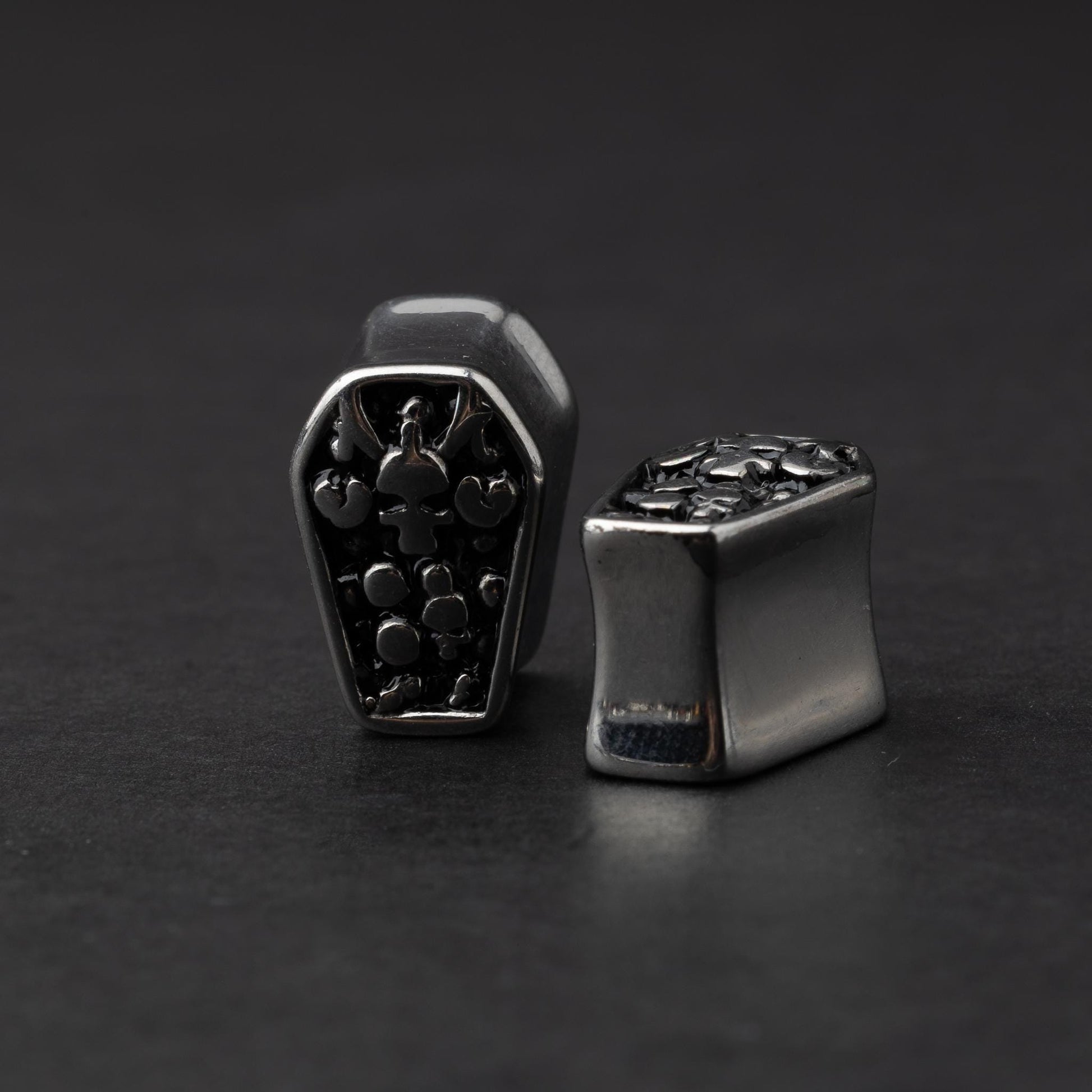 A pair of coffin-shaped stainless steel ear plugs with intricate skeleton designs, displayed on a dark background. In this shot the right one is laying down. 