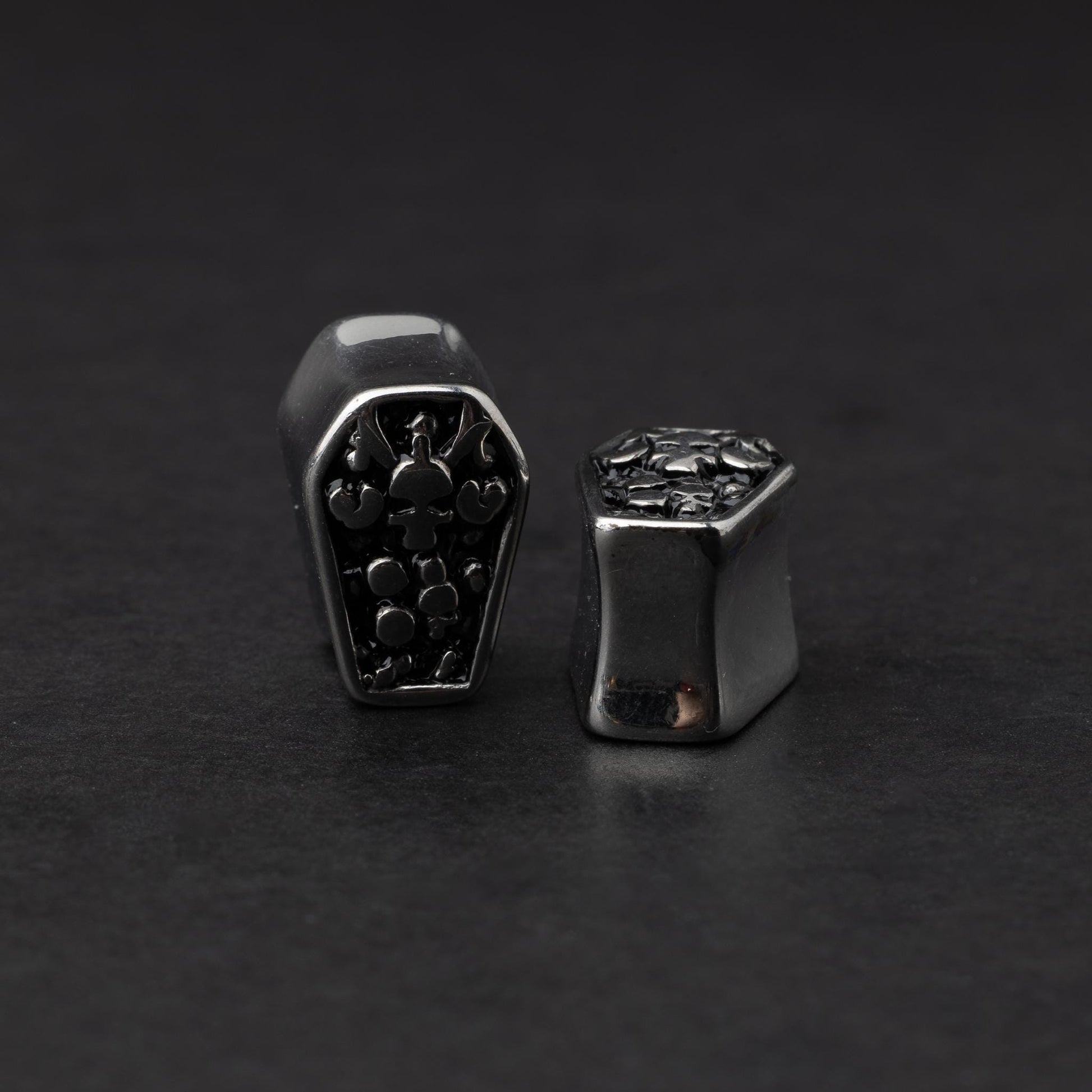 A pair of coffin-shaped stainless steel ear plugs with intricate skeleton designs, displayed on a dark background.