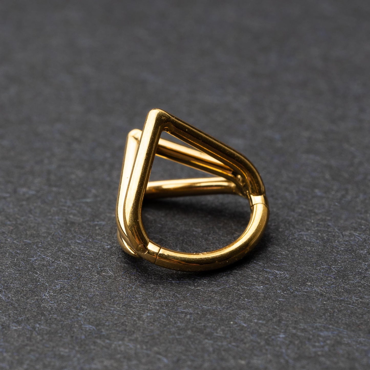 Close-up of a geometric septum clicker in gold, featuring a unique layered triangular design, displayed on a dark background.
