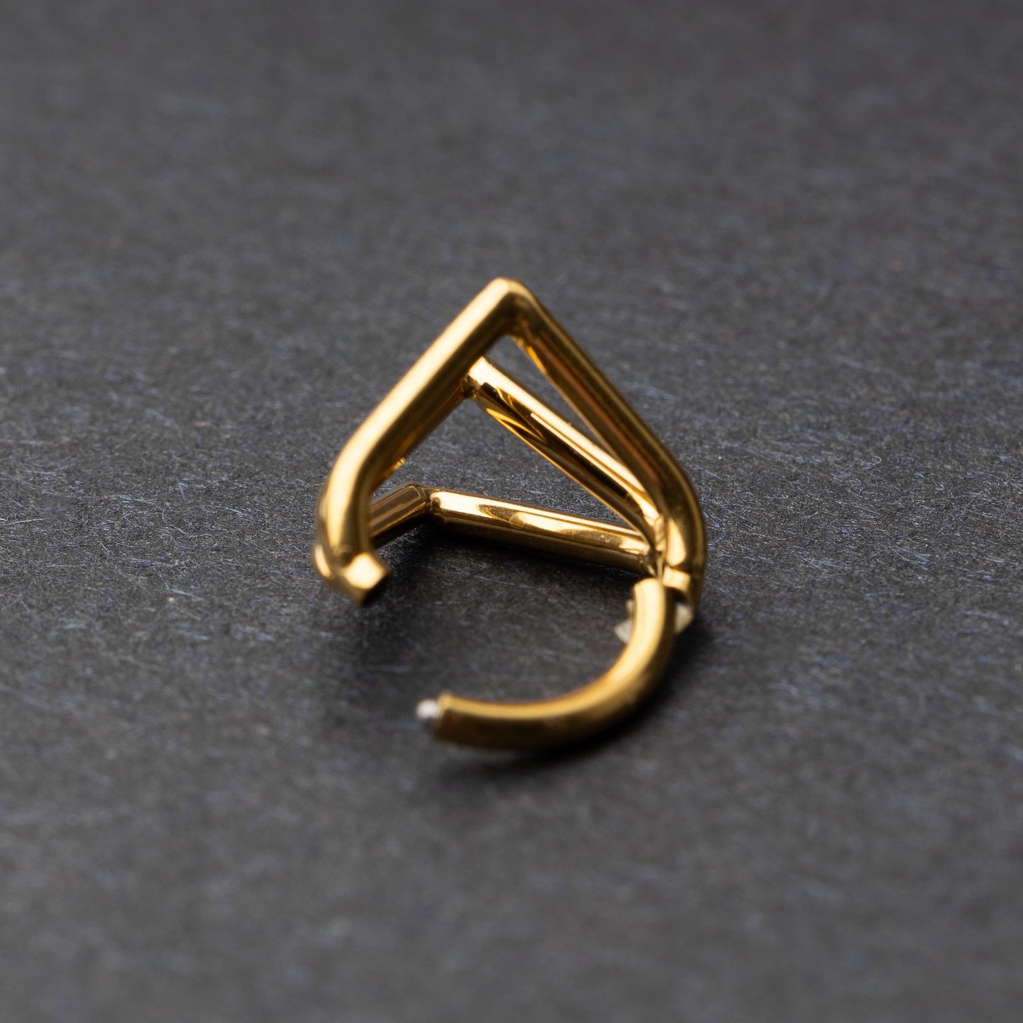 Close-up of a geometric septum clicker in gold, featuring a unique layered triangular design, displayed on a dark background.
