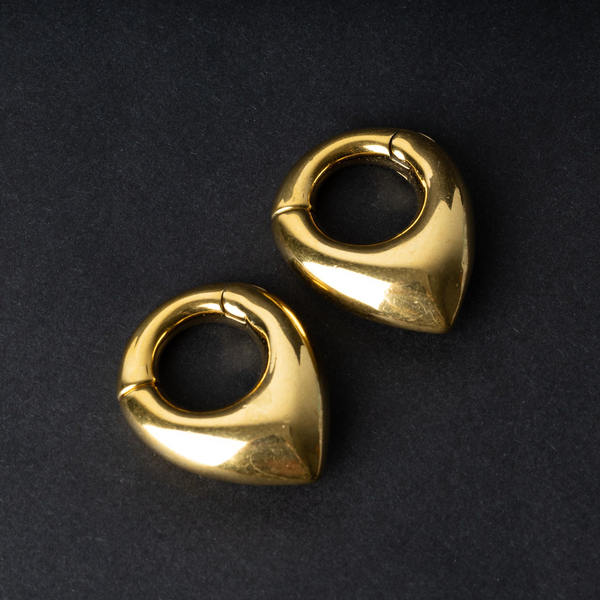 Two teardrop-shaped ear weights, in polished gold, with a seamless hinge closure, displayed on a dark background.
