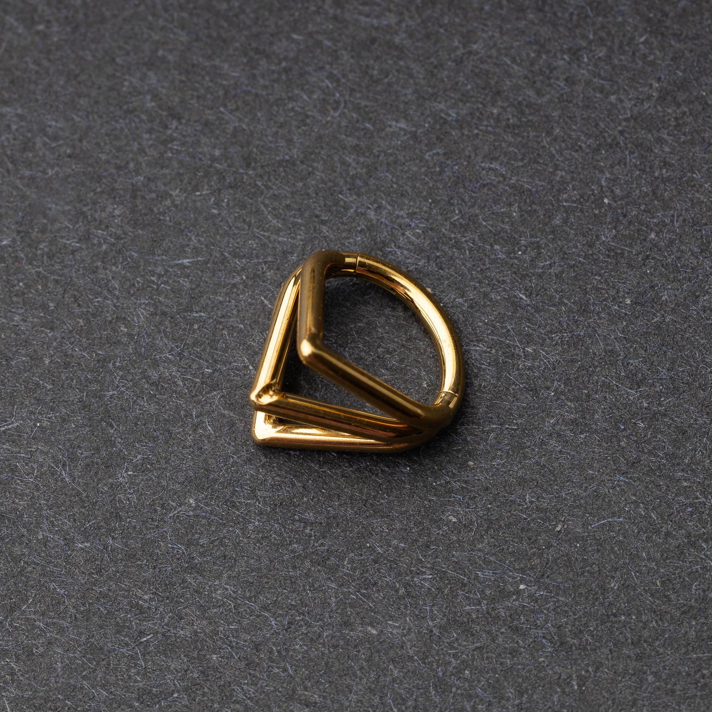 Close-up of a geometric septum clicker in gold, featuring a unique layered triangular design, displayed on a dark background.
