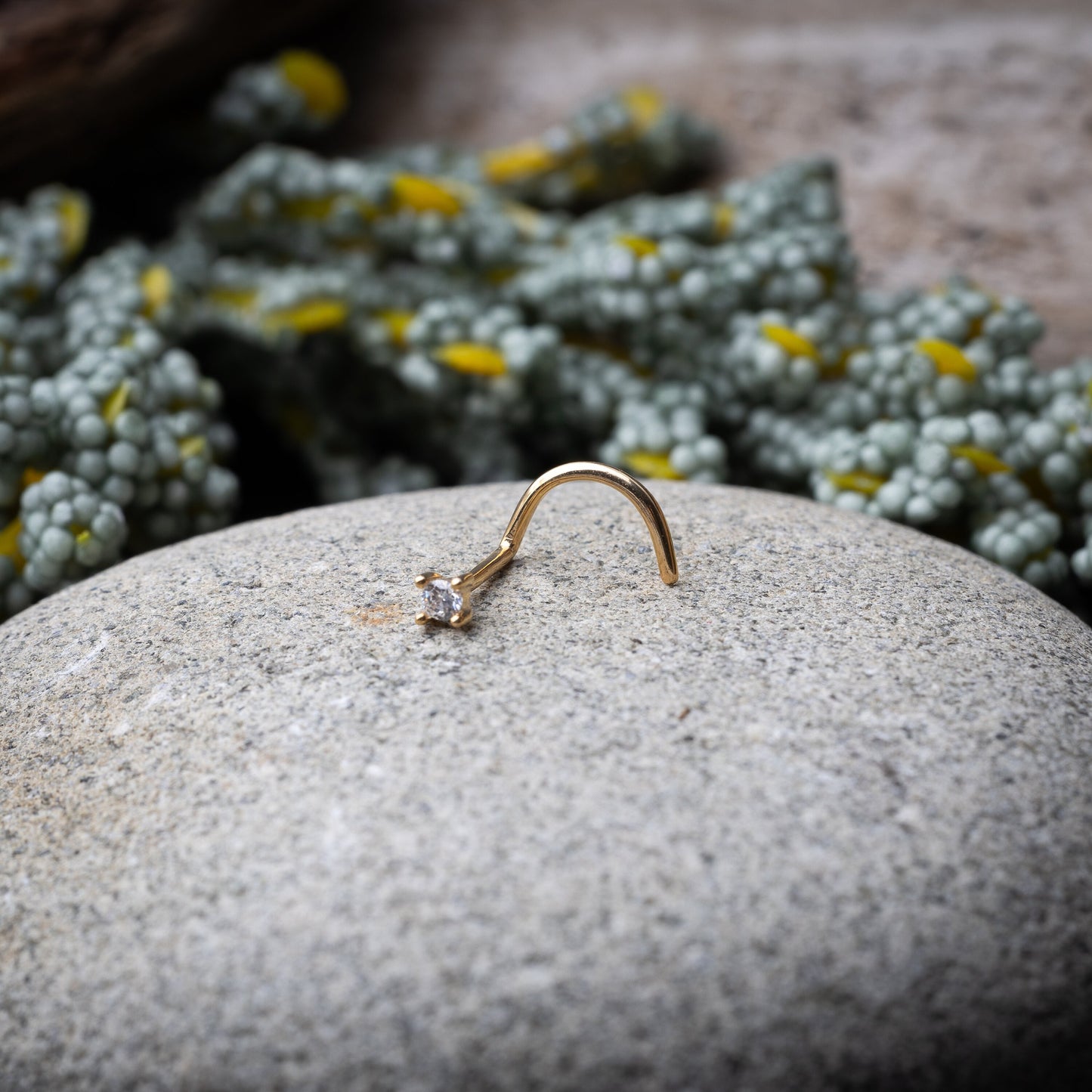 A gold-tone nose stud with a clear prong-set gem, featuring a curved design for secure wear, displayed on a stone background.