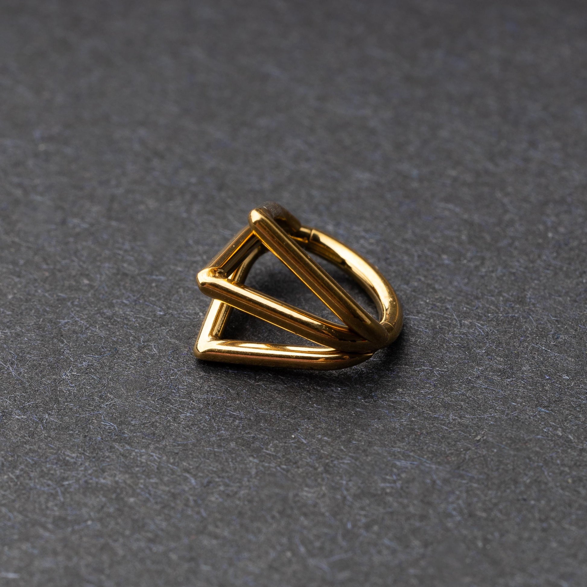 Close-up of a geometric septum clicker in gold, featuring a unique layered triangular design, displayed on a dark background.
