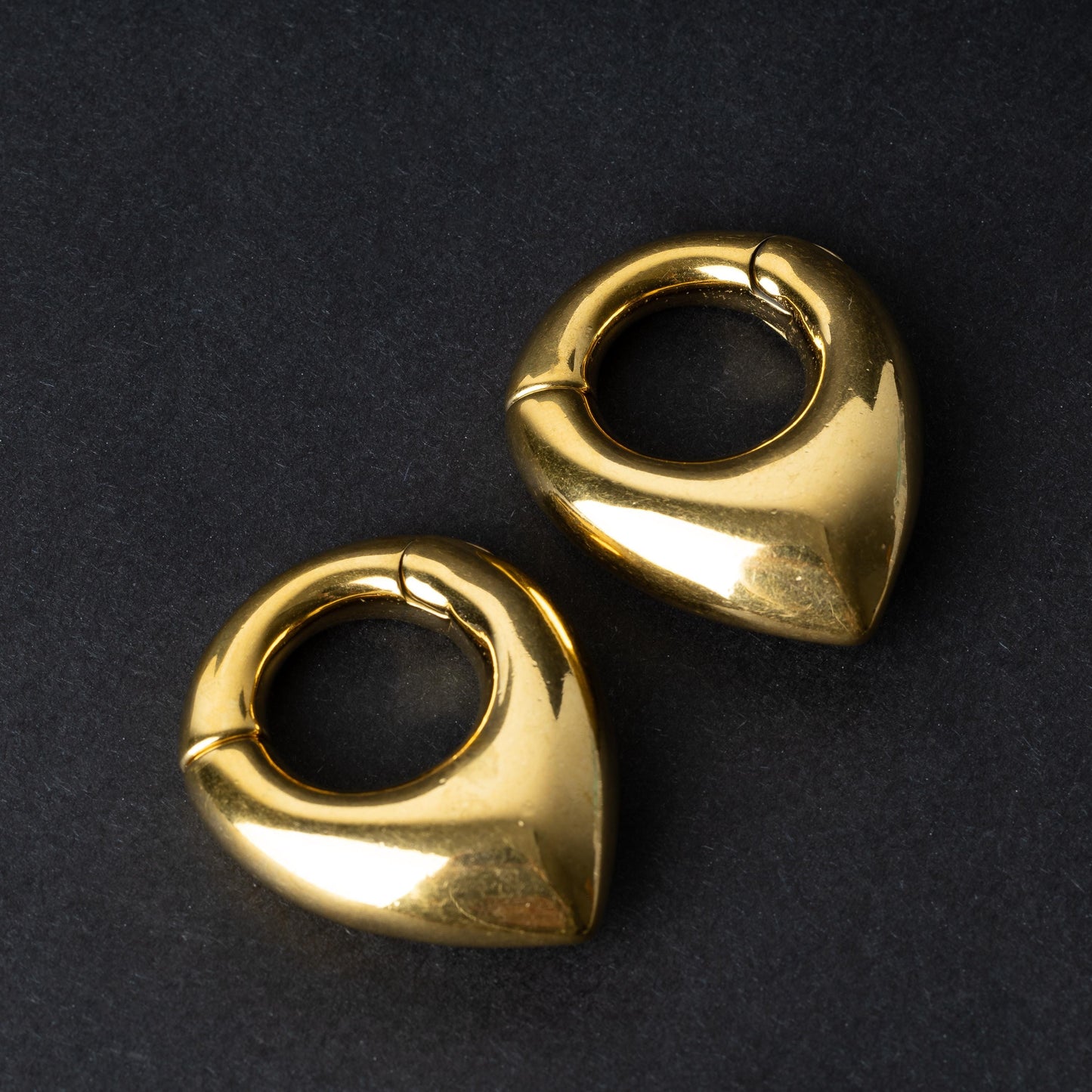 Two teardrop-shaped ear weights, in polished gold, with a seamless hinge closure, displayed on a dark background.

