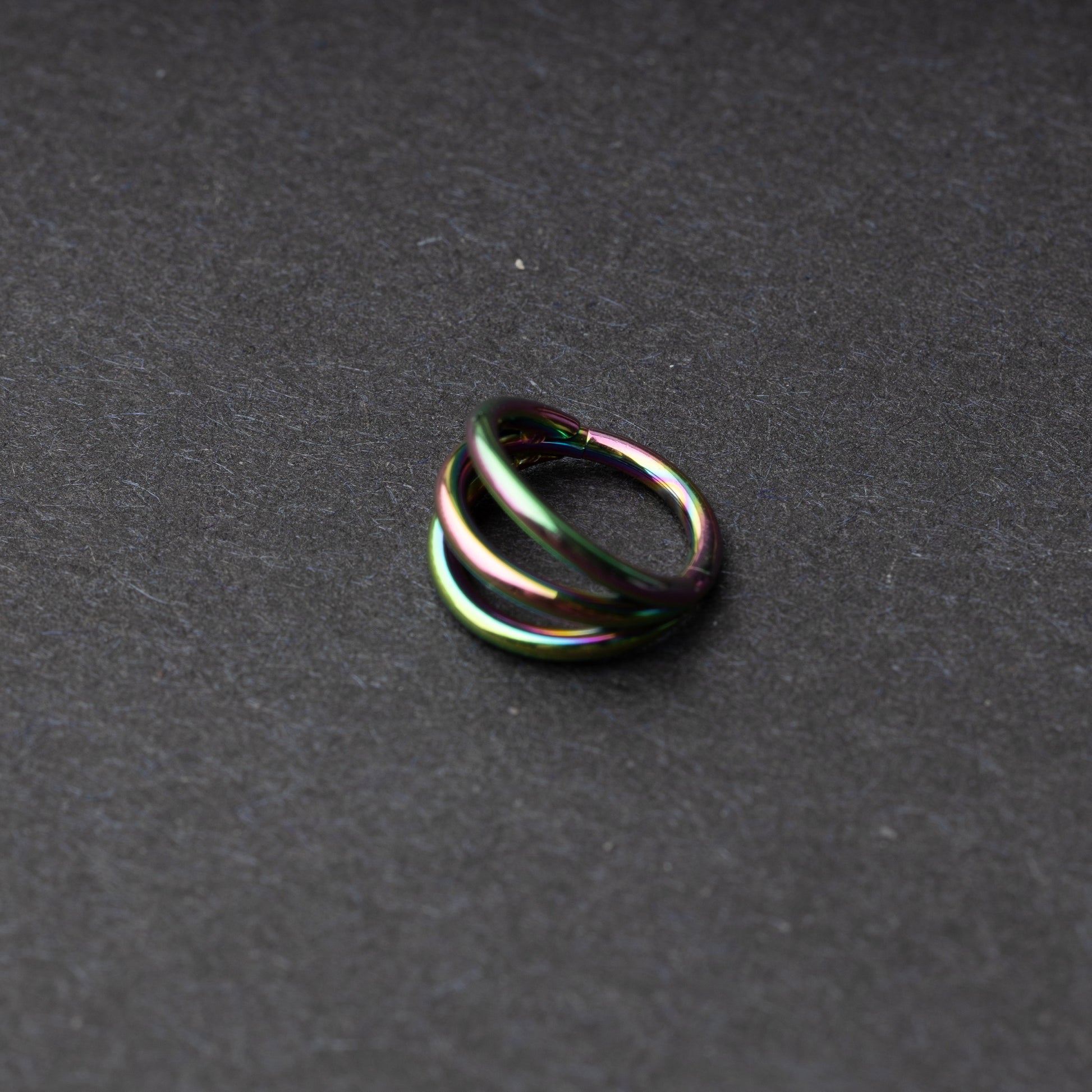 Four triple-band hoop septum clicker ring in rainbow finish, displayed on a dark background.
