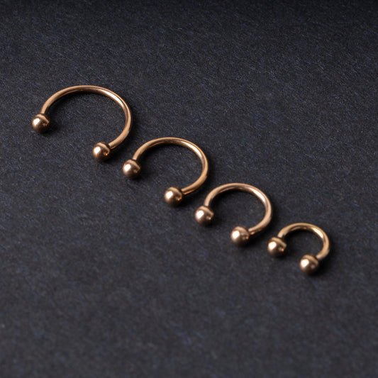 Rose Gold-tone horseshoe rings displayed in a row on a dark background, showcasing various sizes for septum or body piercings.

