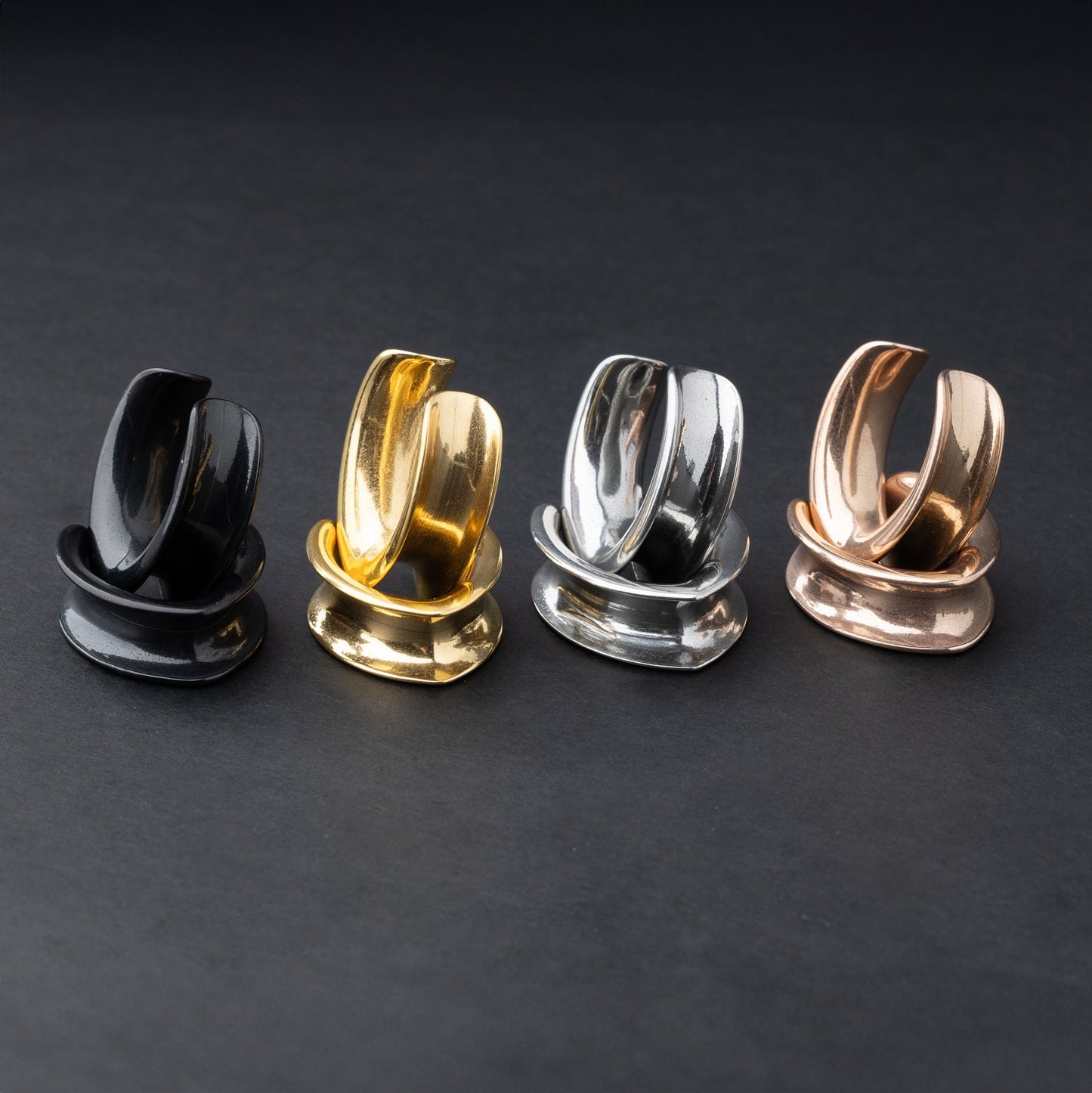 A collection of double-flared metal tunnels for stretched ears in gold, silver, black and rose gold  finish, displayed on a dark background.
