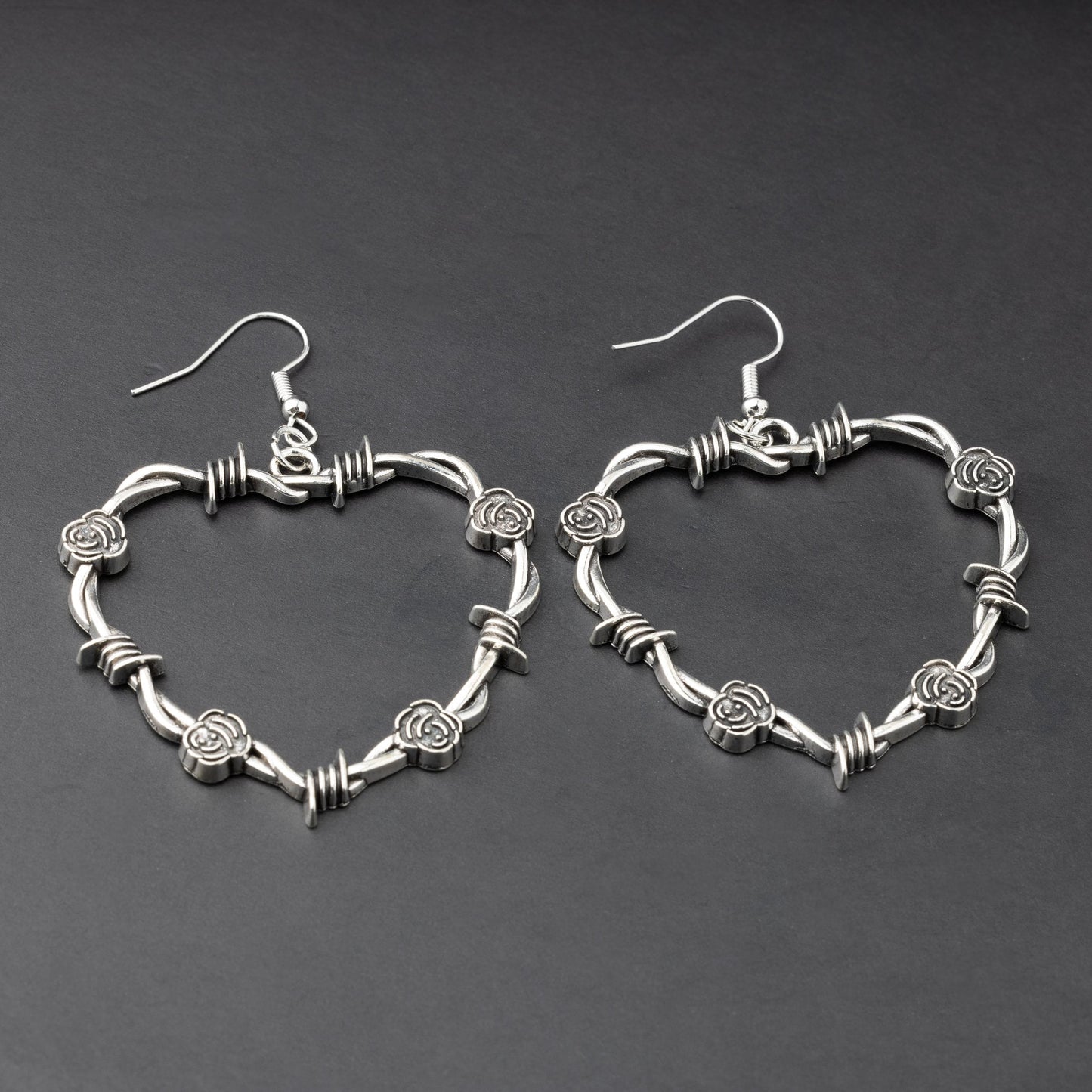 A pair of heart-shaped silver hoop earrings designed with barbed wire detailing, displayed on a dark background.