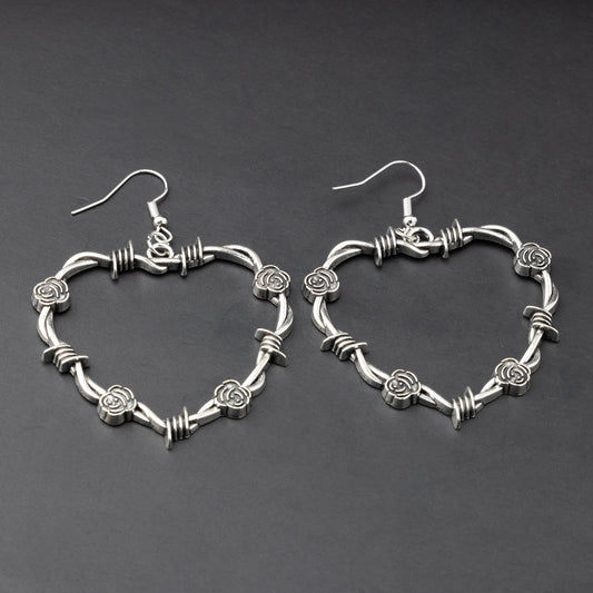 A pair of heart-shaped silver hoop earrings designed with barbed wire detailing, displayed on a dark background.