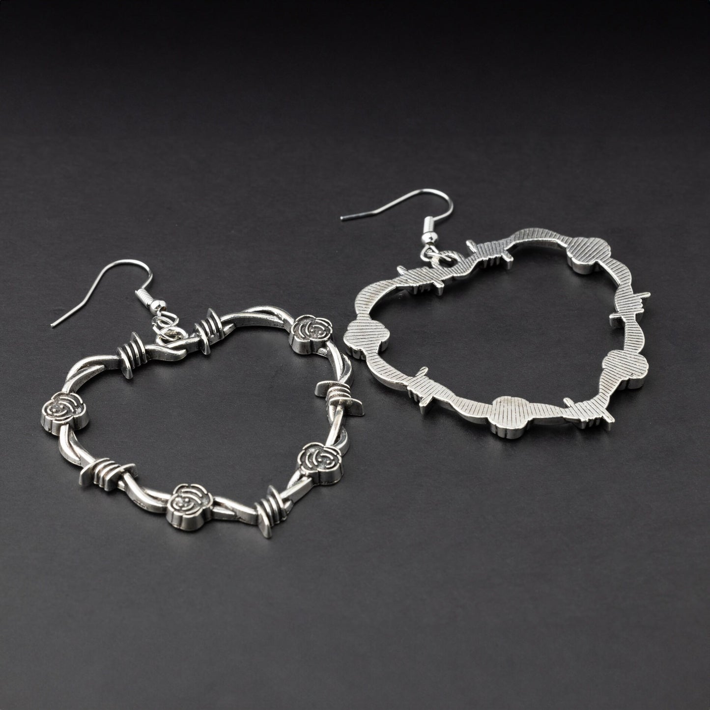 A pair of heart-shaped silver hoop earrings designed with barbed wire detailing, displayed on a dark background.