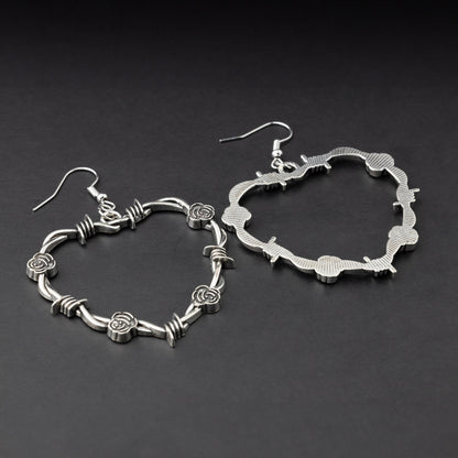 A pair of heart-shaped silver hoop earrings designed with barbed wire detailing, displayed on a dark background.