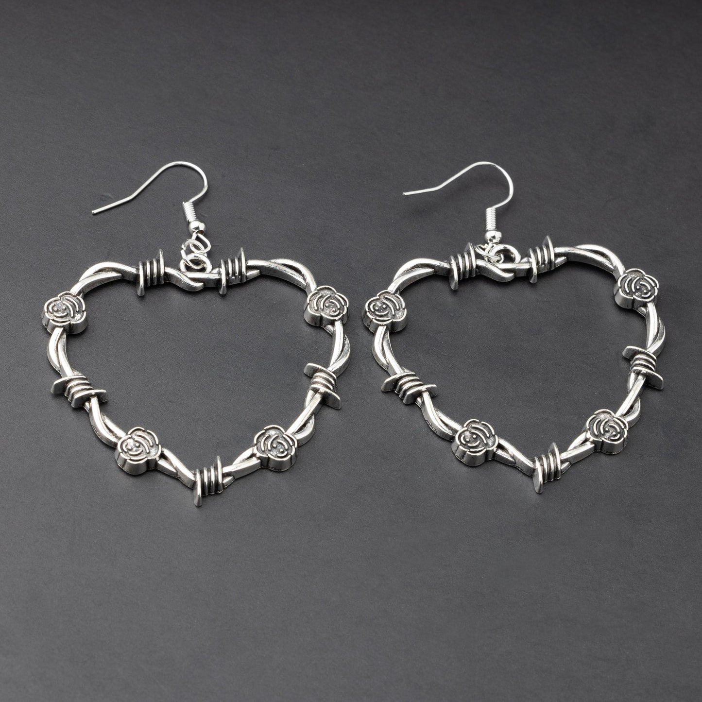 A pair of heart-shaped silver hoop earrings designed with barbed wire detailing, displayed on a dark background.