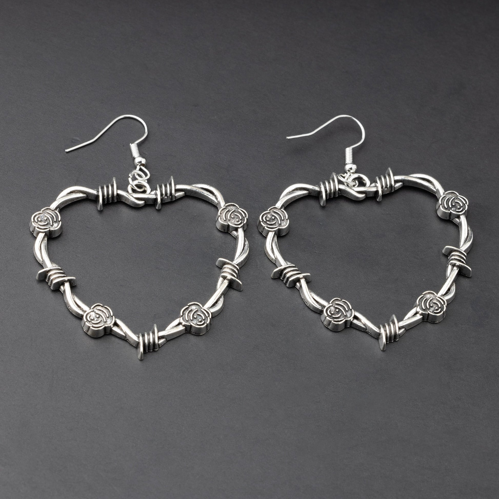 A pair of heart-shaped silver hoop earrings designed with barbed wire detailing, displayed on a dark background.