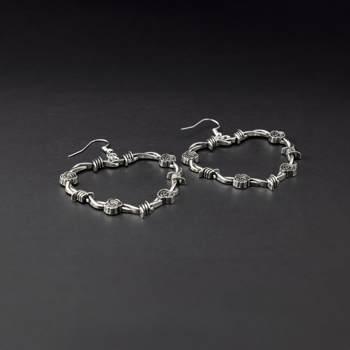 A pair of heart-shaped silver hoop earrings designed with barbed wire detailing, displayed on a dark background.