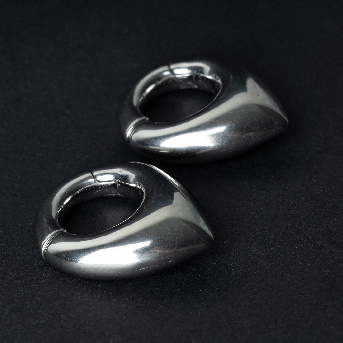 Two teardrop-shaped ear weights, in polished silver, with a seamless hinge closure, displayed on a dark background.

