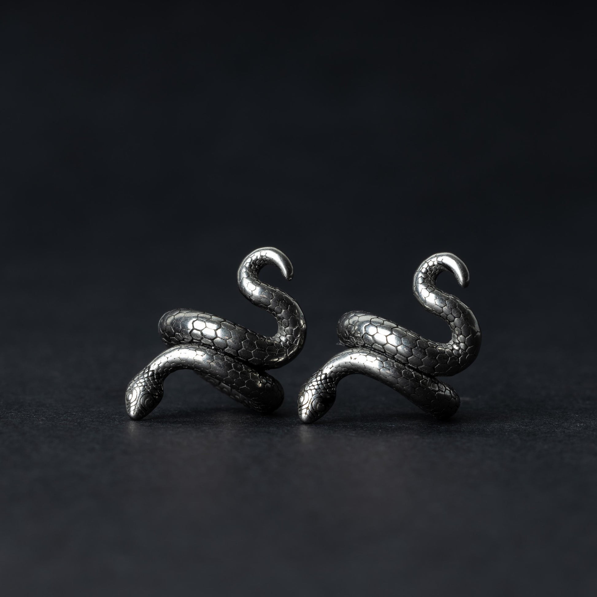 A pair of snake-shaped ear cuffs in silver finish, coiled to wrap around the earlobe, displayed on a dark background.
