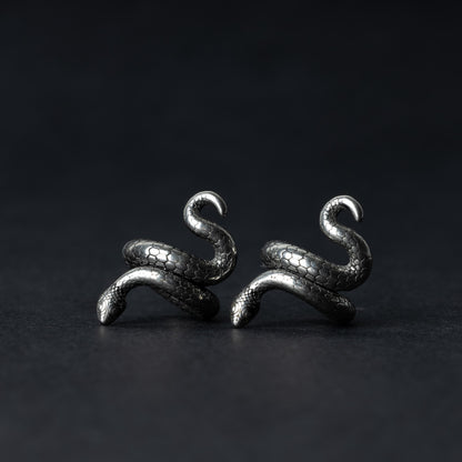 A pair of snake-shaped ear cuffs in silver finish, coiled to wrap around the earlobe, displayed on a dark background.
