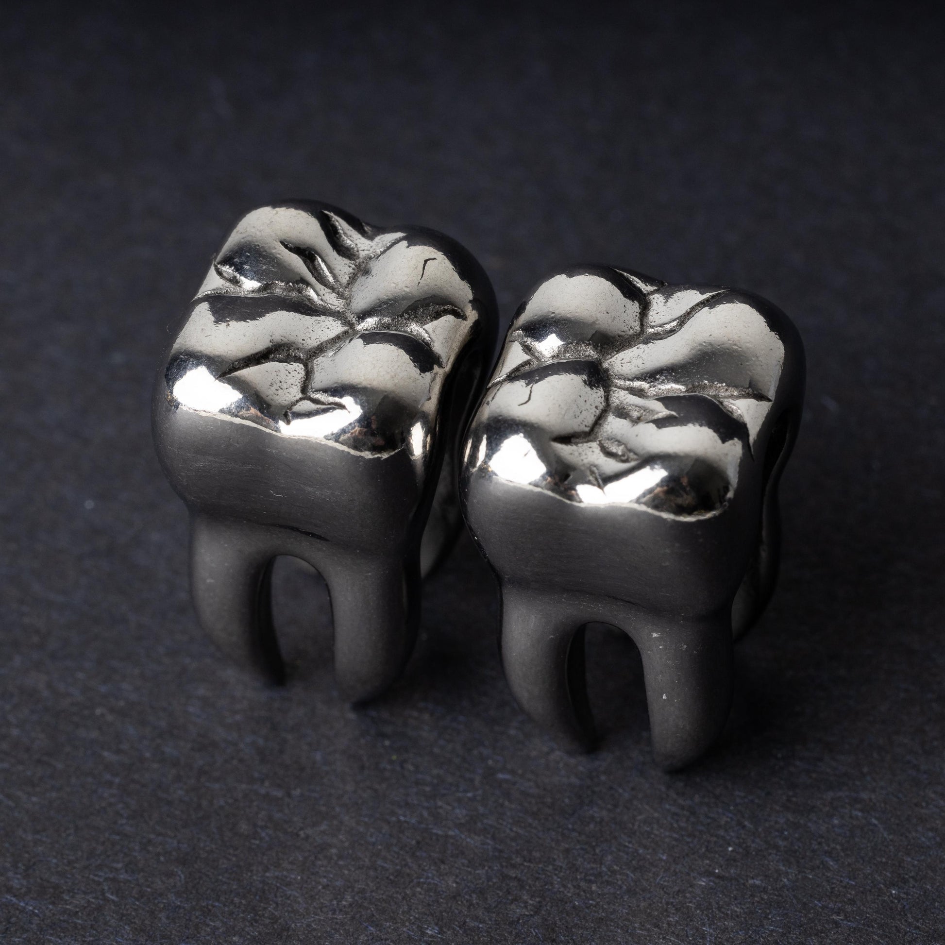 Close-up of silver tooth-shaped ear weights with polished detailing, displayed on a dark background.

