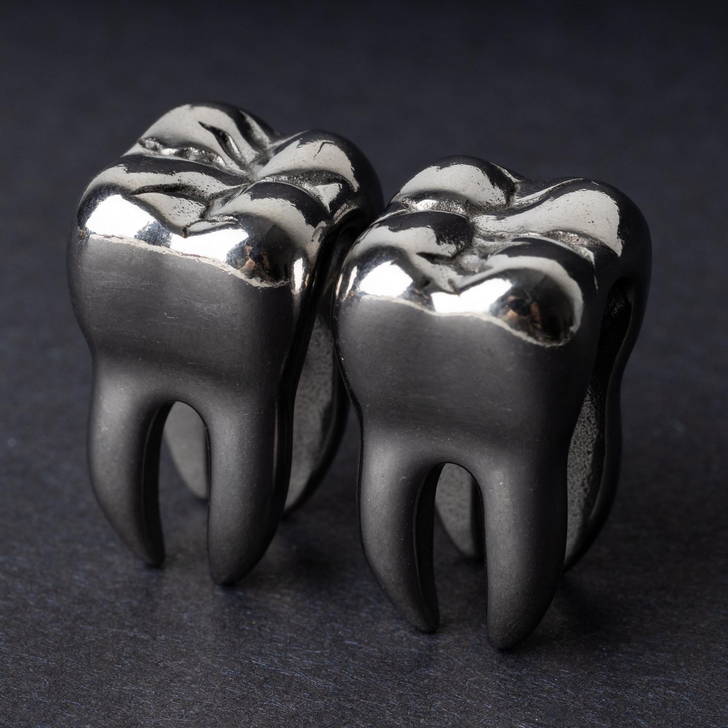 Close-up of silver tooth-shaped ear weights with polished detailing, displayed on a dark background.

