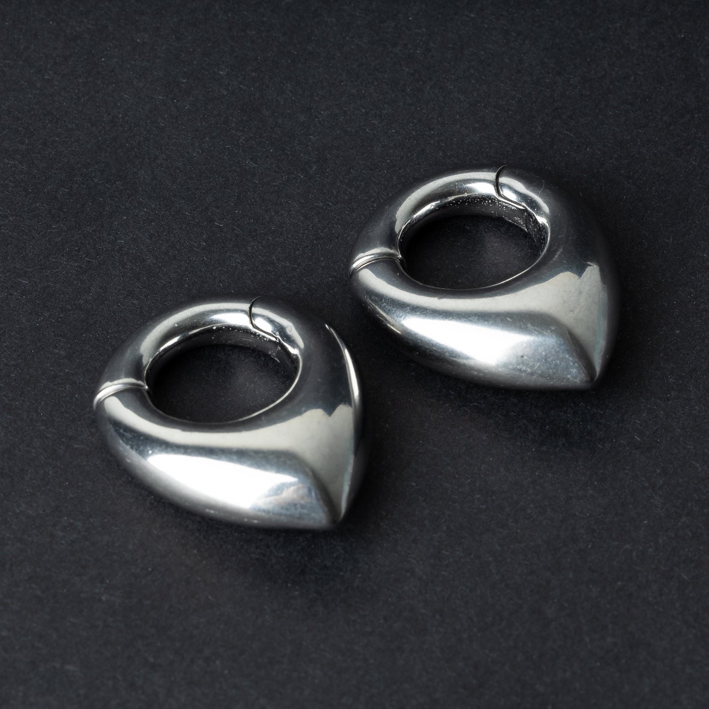 Two teardrop-shaped ear weights, in polished silver, with a seamless hinge closure, displayed on a dark background.

