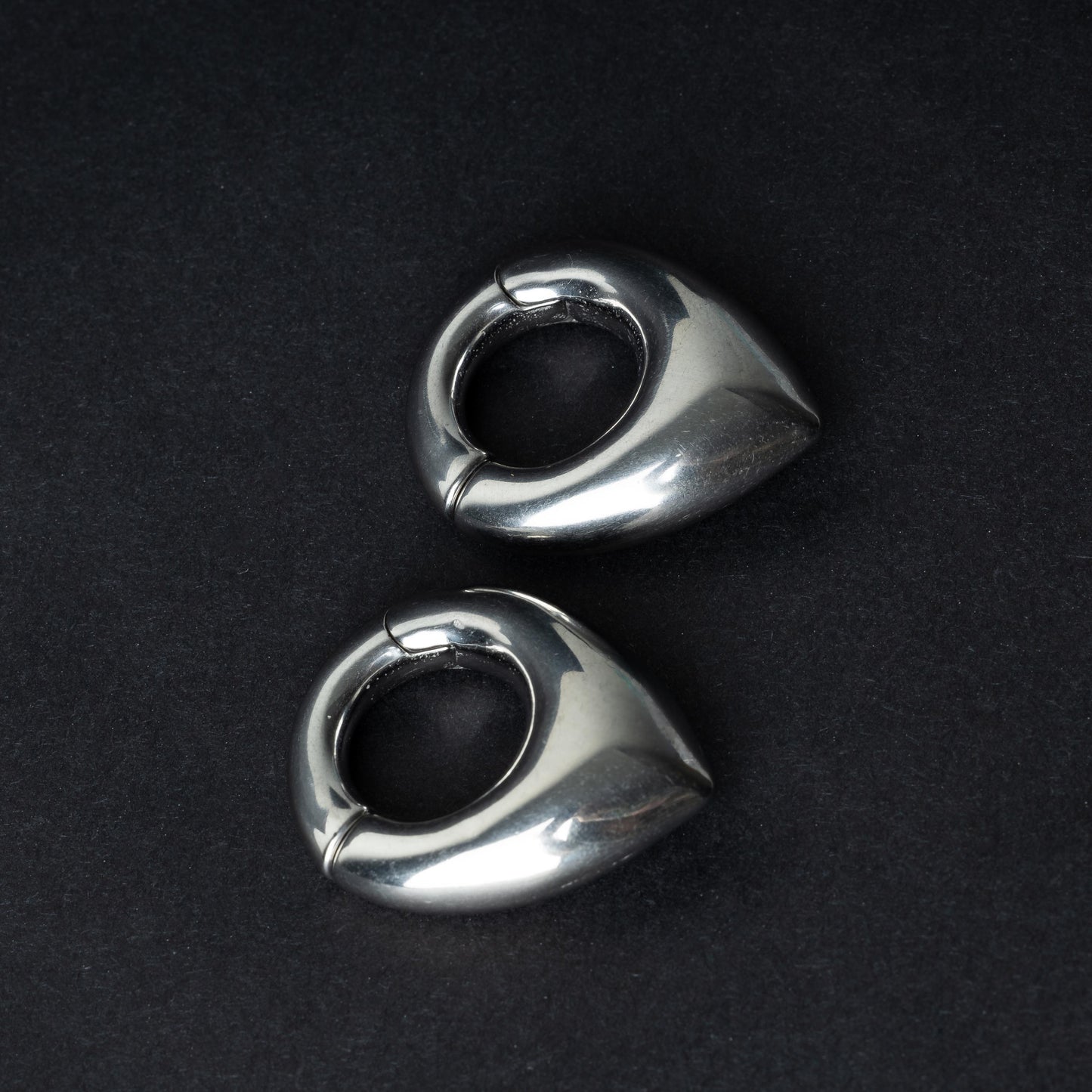 Two teardrop-shaped ear weights, in polished silver, with a seamless hinge closure, displayed on a dark background.
