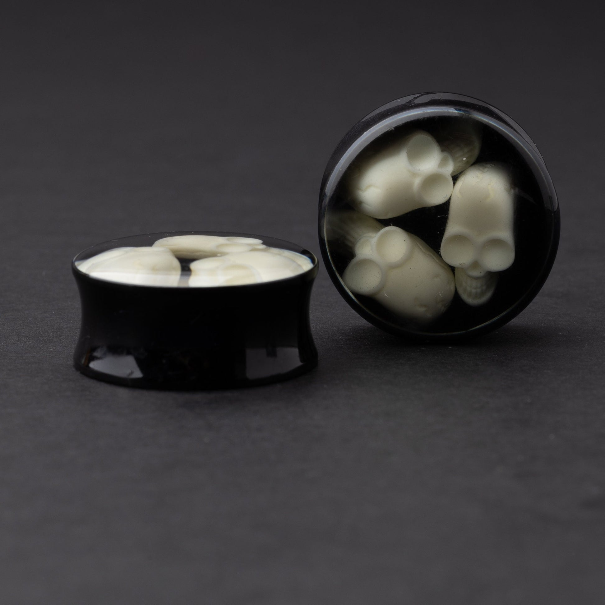 A pair of black double-flared plugs with glow-in-the-dark skulls encased in a polished finish, displayed on a dark background.