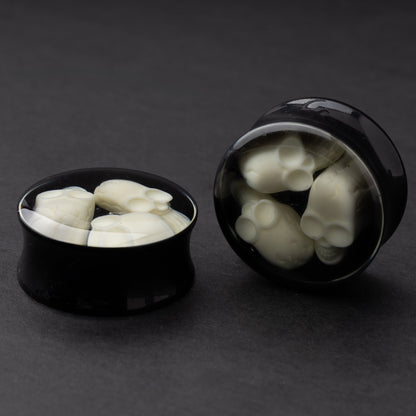 A pair of black double-flared plugs with glow-in-the-dark skulls encased in a polished finish, displayed on a dark background.