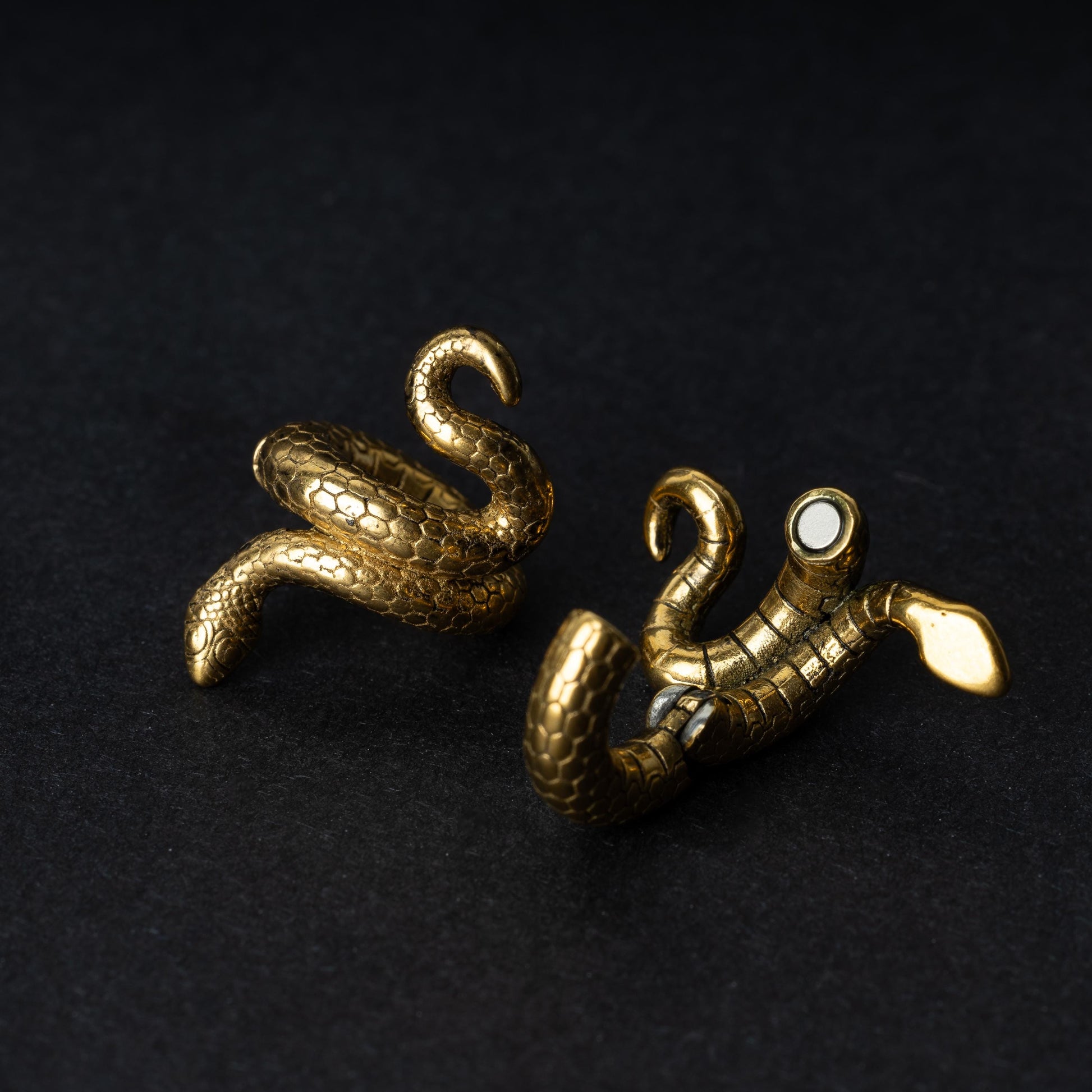 A pair of snake-shaped ear cuffs in gold finish, coiled to wrap around the earlobe, displayed on a dark background.

