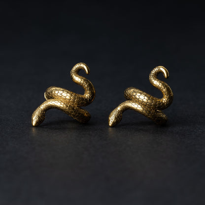 A pair of snake-shaped ear cuffs in gold finish, coiled to wrap around the earlobe, displayed on a dark background.
