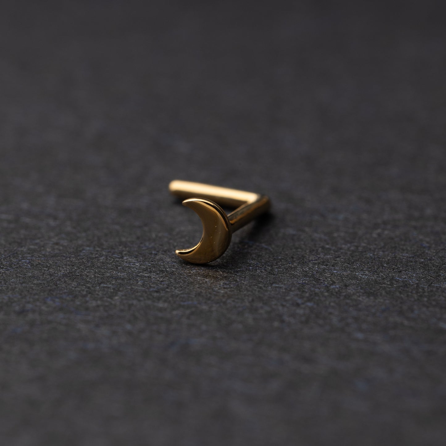 A gold-tone crescent moon nose stud with a curved design for secure and comfortable wear, displayed on a dark background.