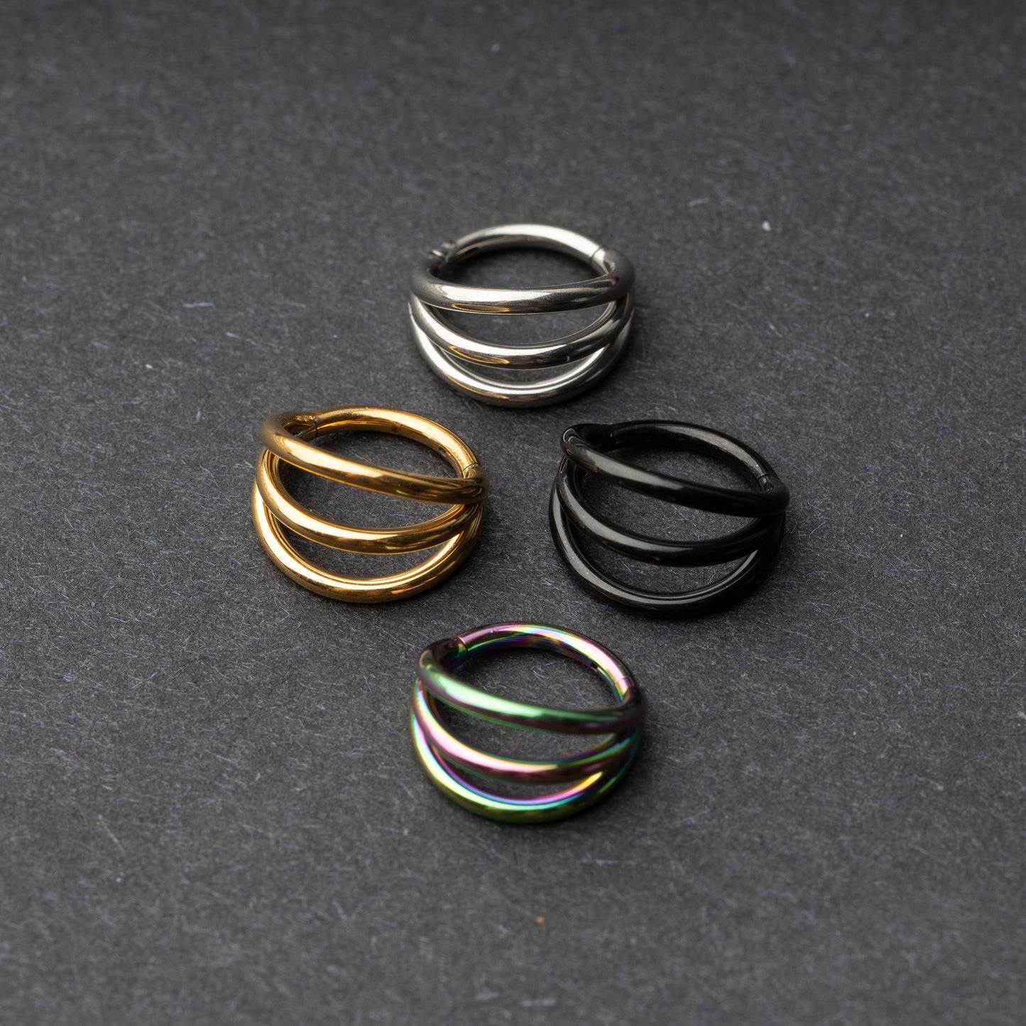 Four triple-band hoop septum clicker rings in gold, silver, black, and rainbow finishes displayed on a dark background.
