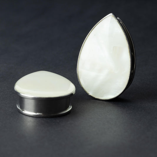 A pair of teardrop-shaped plugs with a pearlescent shell inlay, framed by stainless steel, resting on a dark background.

