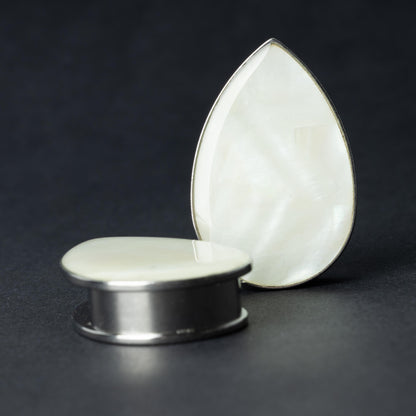 A pair of teardrop-shaped plugs with a pearlescent shell inlay, framed by stainless steel, resting on a dark background.
