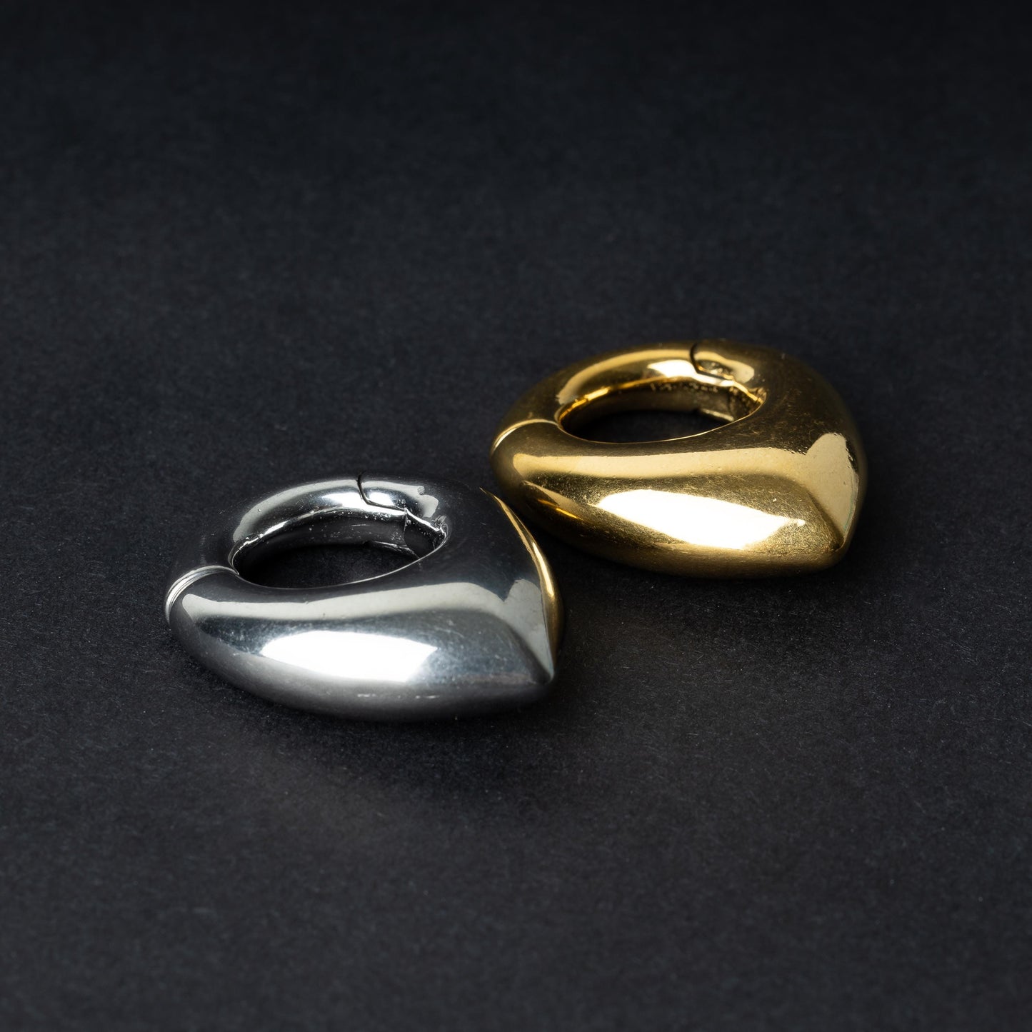 Two teardrop-shaped ear weights, one in polished silver and the other in polished gold, with a seamless hinge closure, displayed on a dark background.

