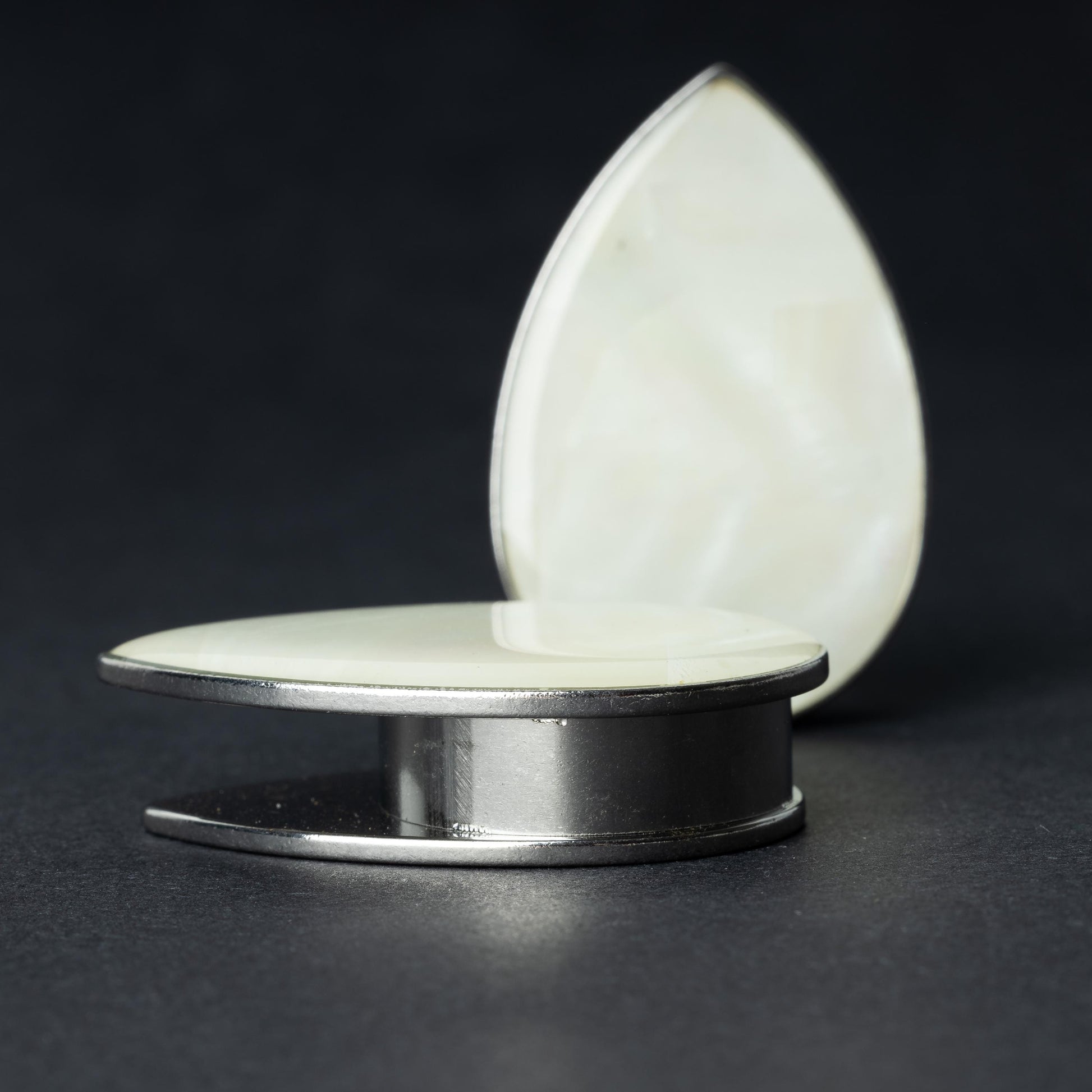 A pair of teardrop-shaped plugs with a pearlescent shell inlay, framed by stainless steel, resting on a dark background.
