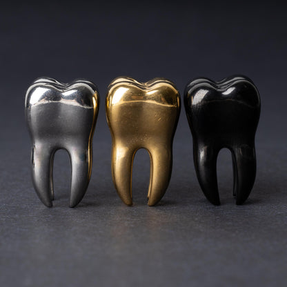 Close-up of gold silver and black tooth-shaped ear weights with polished detailing, displayed on a dark background.

