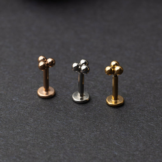 Close-up of three minimalist ball cluster labret studs in rose gold, silver, and gold finishes, displayed on a dark background.

