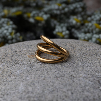 Four triple-band hoop septum clicker ring in gold finish, displayed on a rocky background.
