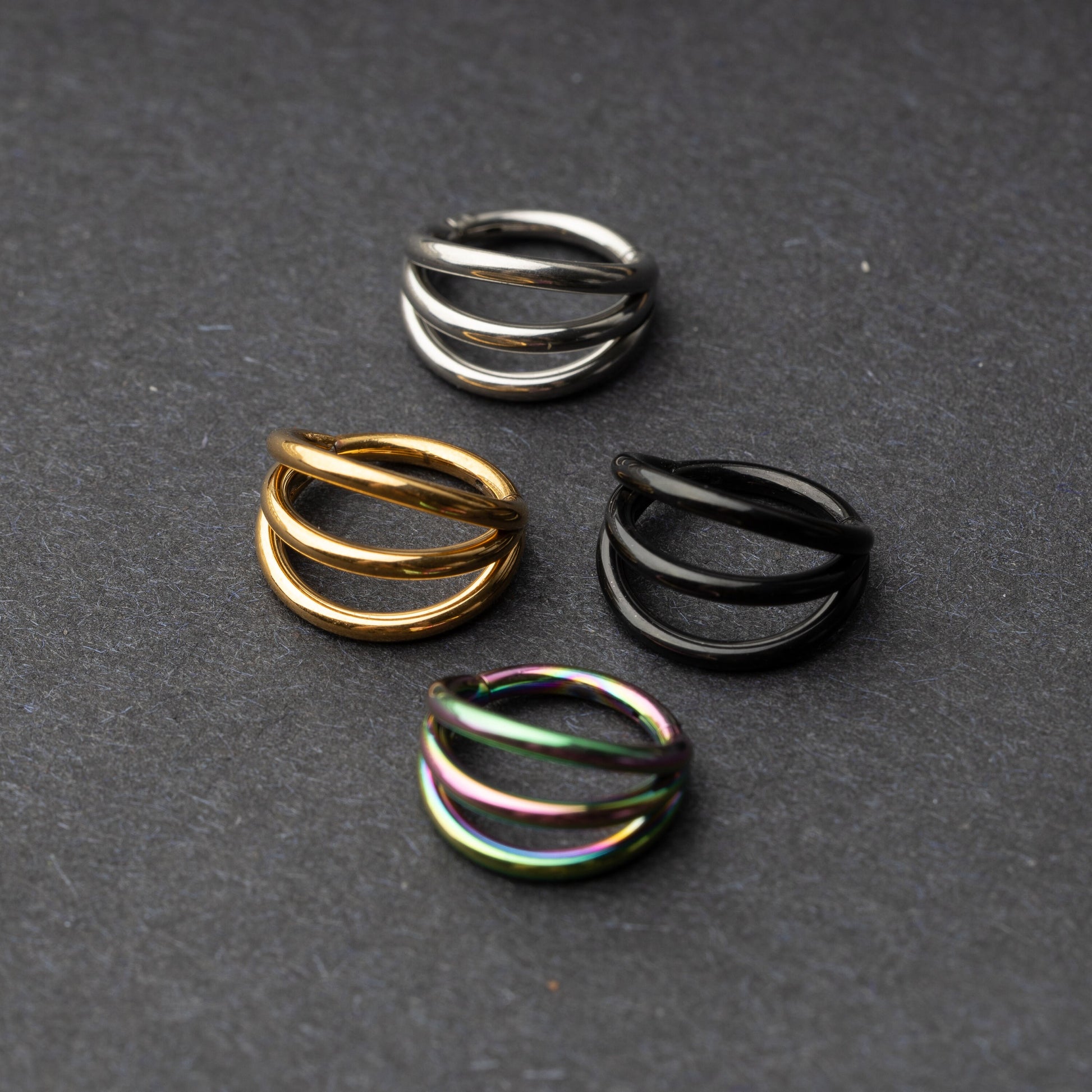 Four triple-band hoop septum clicker rings in gold, silver, black, and rainbow finishes displayed on a dark background.

