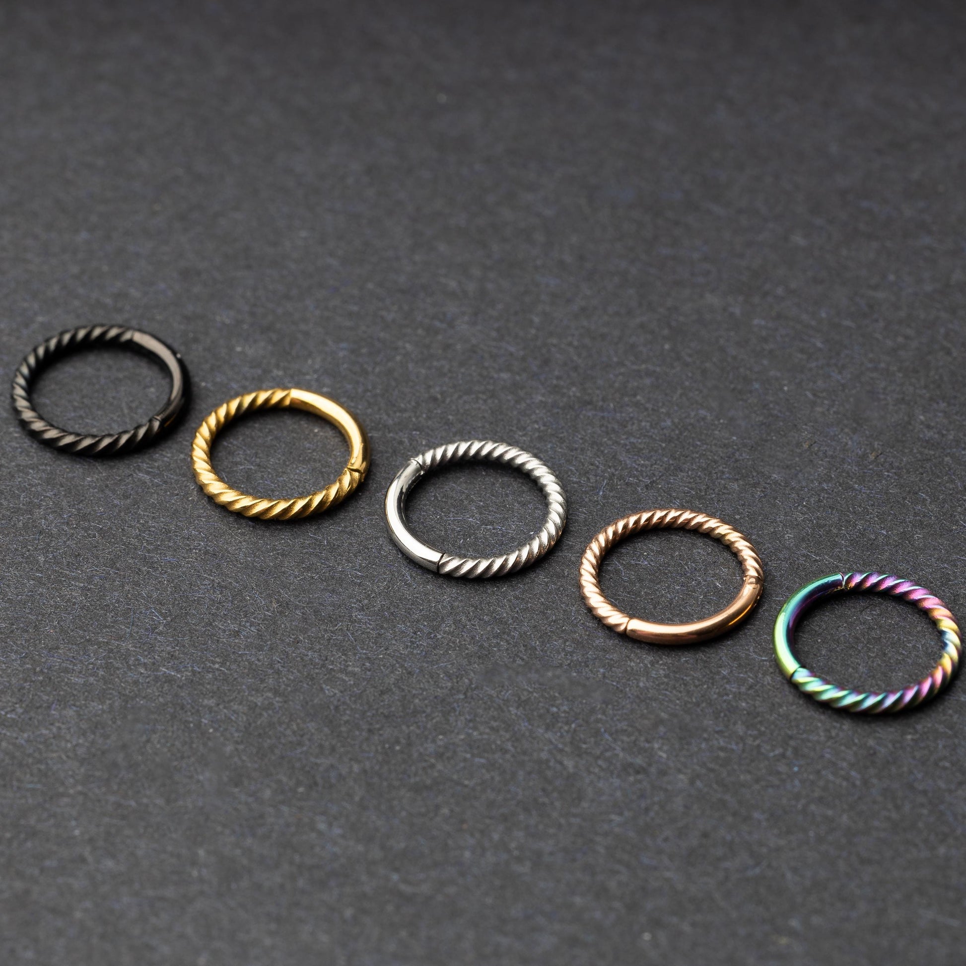 Twisted seamless hoop rings in black, gold, silver, rose gold, and rainbow finishes displayed on a dark background, highlighting their elegant and textured design.