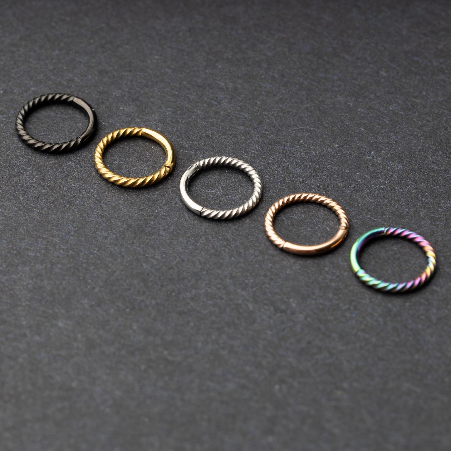 Twisted seamless hoop rings in black, gold, silver, rose gold, and rainbow finishes displayed on a dark background, highlighting their elegant and textured design.