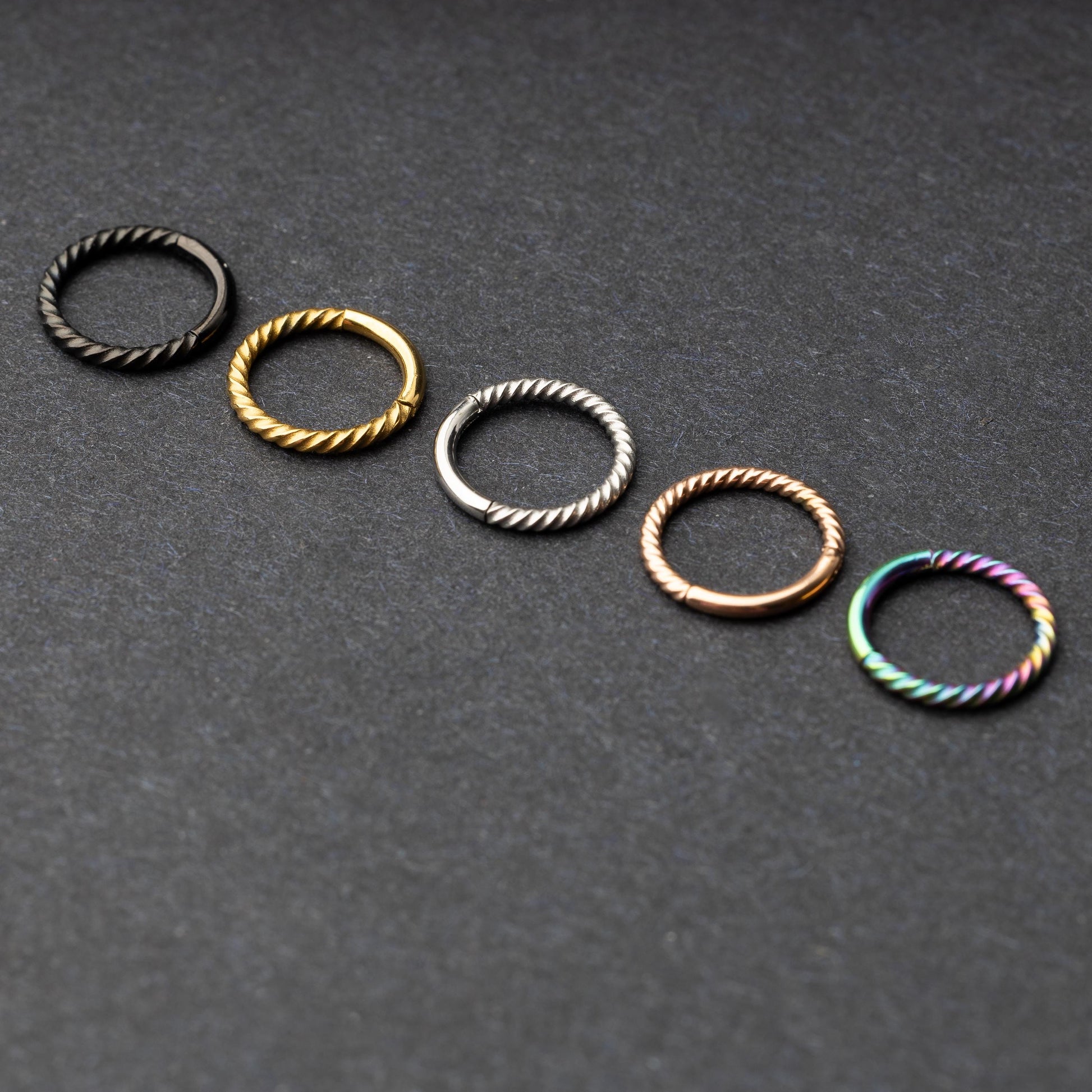 Twisted seamless hoop rings in black, gold, silver, rose gold, and rainbow finishes displayed on a dark background, highlighting their elegant and textured design.