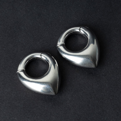 Two teardrop-shaped ear weights, in polished silver, with a seamless hinge closure, displayed on a dark background.
