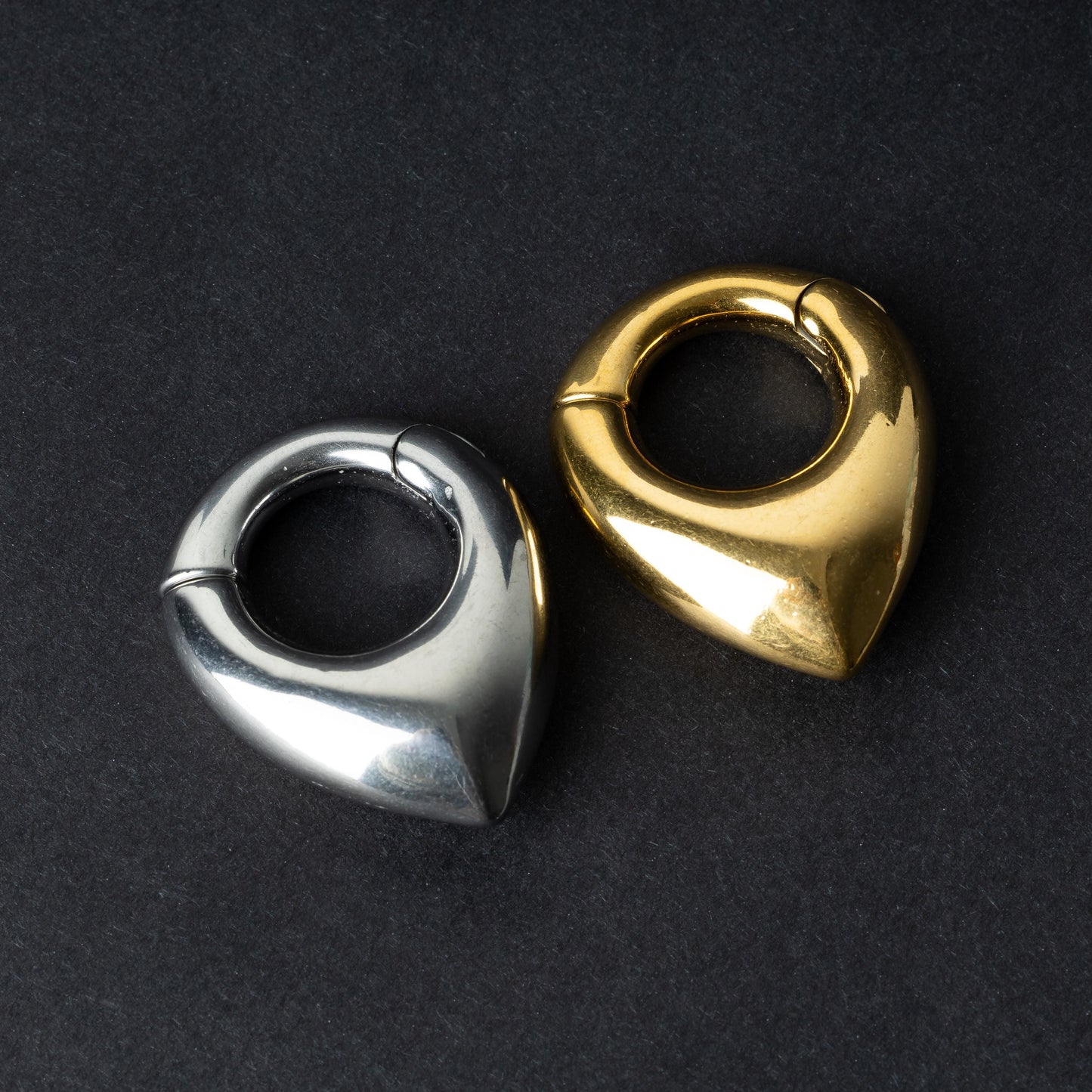 Two teardrop-shaped ear weights, one in polished silver and the other in polished gold, with a seamless hinge closure, displayed on a dark background.

